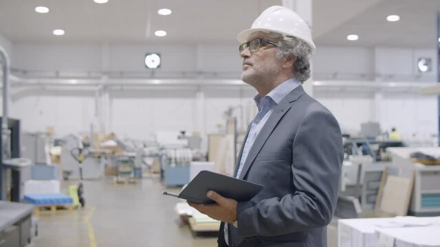 Mature engineer checking factory. Man in helmet and business suit looking around on plant floor, using tablet, looking at camera and smiling. Static shot. Industry and production concept
