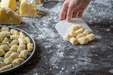 Gnocchi preparation steps: Woman's hand picking up gluten free handmade mashed potato gnocchi using a scraper. Flour sprinkled on a dark table. Creating authentic fresh pasta dough for Italian dish