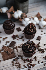 Several muffins or cupcakes with chocolate shaped cream at white table. Festive candle burns on a chocolate cake. There are chocolate chips scattered nearby
