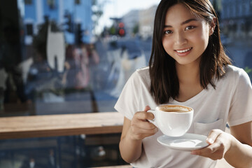 Close-up portrait of young asian woman holding cup of cappuccino in restaurant. Girl drinking coffee in cafe near window and smiling at camera