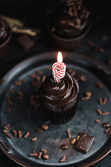Several muffins or cupcakes with chocolate shaped cream at black table. Festive candle burns on a chocolate cake.