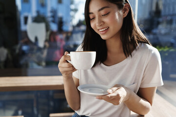 Beautiful asian girl drinking coffee in cafe by the window, looking at cup of cappuccino and smiling on summer day