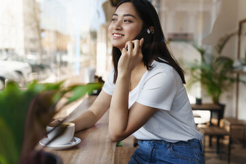 Happy asian candid woman sitting in cafe near the window, looking at street while talking on phone with a friend, smiling and drinking coffee