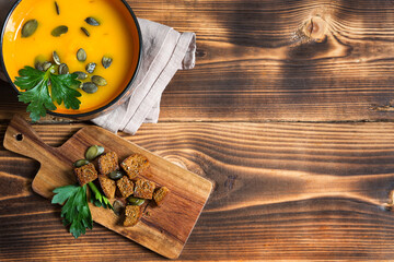 Pumpkin soup in a bowl with seeds and parsley on a wooden background. Crackers with spices on the board. Copy space
