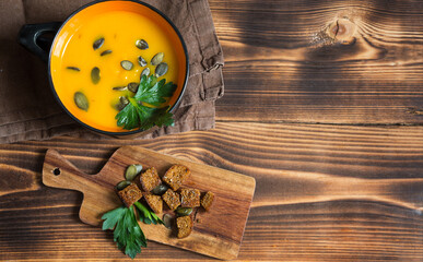 Obraz na płótnie Canvas Pumpkin soup in a bowl with seeds and parsley on a wooden background. Crackers with spices on the board. Copy space
