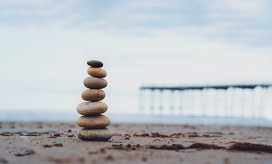 Pebble tower by the seaside with blurry pier down to the sea, Stack of zen rock stones on the sand, Stones pyramid on the beach symbolizing, stability, harmony, balance with shallow depth of field.