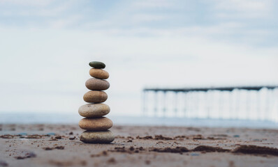 Fototapeta na wymiar Pebble tower by the seaside with blurry pier down to the sea, Stack of zen rock stones on the sand, Stones pyramid on the beach symbolizing, stability, harmony, balance with shallow depth of field.