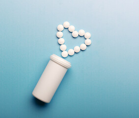 Open container with pills scattered in the heart shape on  blue background.