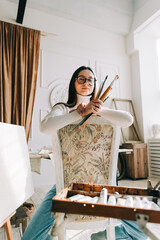 Portrait of beautiful smiling woman artist, sitting on a chair and holding paintbrushes, working on a home studio.