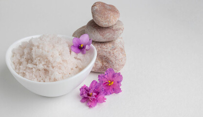 Pink coarse salt in a white bowl stands on a white background next to pink stones and pink violet flowers