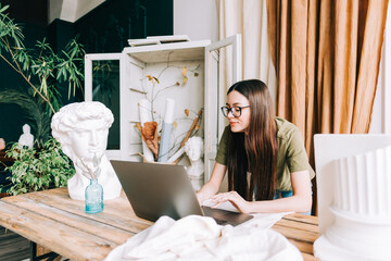 Young caucasian woman in eyeglasses working on laptop computer in home art studio.