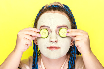 Woman with blue afro braids face in a frozen mask of green clay with slices of cucumbers in front of her eyes on a yellow background.