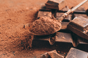Brown cocoa powder in the spoon and chopped chocolate cubes, on dark background