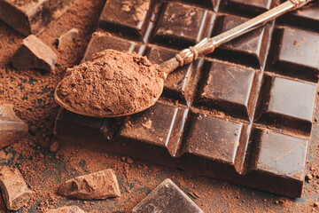 Brown cocoa powder in the spoon, chocolate bar and chopped chocolate cubes, on dark background