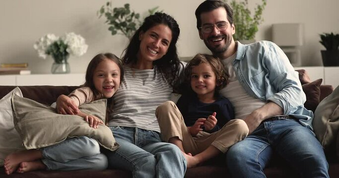 Married couple their little son and daughter sit on couch smile look at camera, parents and kids tickling each other laughing feels carefree enjoy playtime at modern home. Happy family games concept