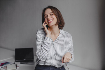 Portrait of beautiful woman in classic blouse talking on phone in white office with laptop on table