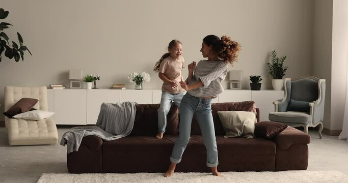 Funny mom and little daughter dancing fooling together in cozy warm living room during active weekend at home. Mother or babysitter holding hands of kid girl swings whirls her laughing enjoy playtime