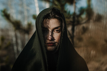 Artistic portrait of a young woman with a scarf over head, shadows on face, deep, dramatic look, in a forest background