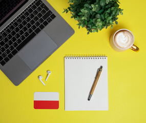 online Polish language courses. Layout of laptop, headphones, Polish flag, notepad with pen, cappuccino coffee and plant on yellow background