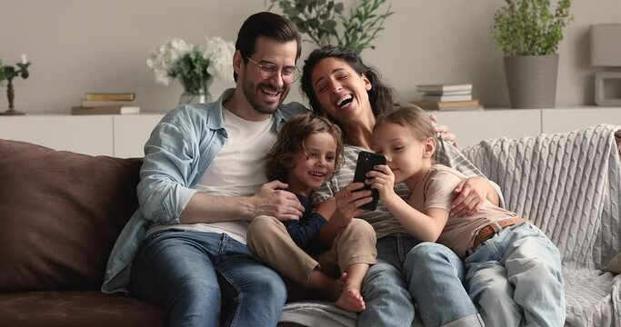 Young couple with son daughter relaxing on couch in living room talking having fun laughing, family spend free time together at home use smartphone cool mobile app. Weekend, online amusements concept