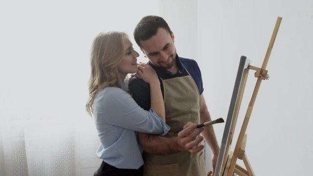 Young male artist with apron paints on canvas drawing picture with his girlfriend in art studio.