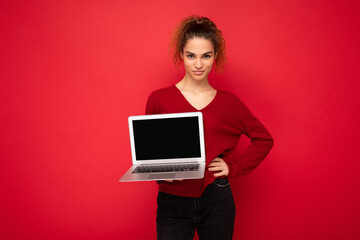 Charming young brunette curly lady looking at camera with confident sight holding computer laptop with empty copy space mock up keeps arm at hip wearing red sweater isolated on red wall background