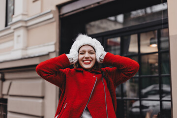 Close-up portrait of joyful woman with red lipstick, laughing with closed eyes. Girl in warm coat, hat and mittens touches her head