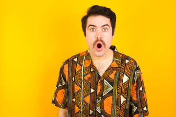 young Caucasian man wearing printed shirt against yellow wall having stunned and shocked look, with mouth open and jaw dropped exclaiming: Wow, I can't believe this. Surprise and shock