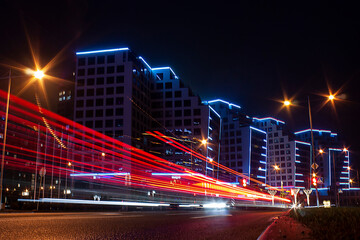 Fototapeta na wymiar Horizontal long exposure photo with night city street, modern buildings along the road with blue illumination and red tail light trails of vehicle