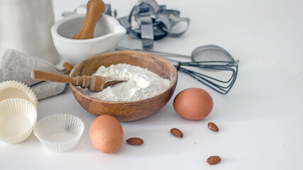 Fototapeta na wymiar Homemade baking ingredients and accessories. A jar of sugar, a cup of flour, eggs, a whisk, a cake pan, a napkin, nuts on a white table. Home hobbies authentic cooking concept, close-up, copy space