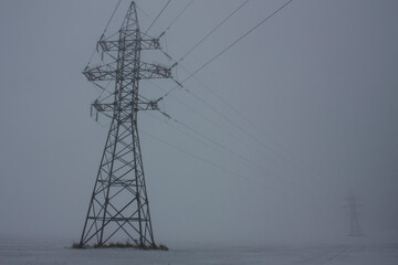 Horizontal outdoors dim photography with a steel electricity high voltage power line support in the fog on the field during overcast winter day