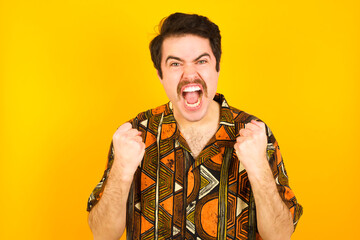 young Caucasian man wearing printed shirt against yellow wall rejoicing success and victory clenching his fists with joy being happy to achieve her aim and goals. Positive emotions, feelings.