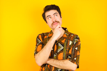 Portrait of thoughtful young Caucasian man wearing printed shirt against yellow wall keeps hand under chin, looks away trying to remember something or listens something with interest. Youth concept.