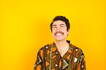 Portrait of mysterious young Caucasian man wearing printed shirt against yellow wall looking up with enigmatic smile. Advertisement concept.