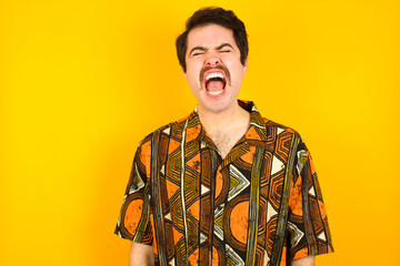 Stressful young Caucasian man wearing printed shirt against yellow wall screams in panic, closes eyes in terror, keeps hands on head, finds out terrified news, can't believe it.