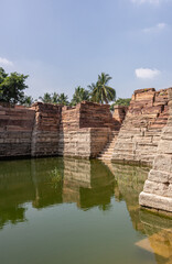 Fototapeta na wymiar Aihole, Karnataka, India - November 7, 2013: Weeds grow in greenish water of Chakra Gudi tank with brown stone walls on sides and one stairway leading into it. Shot at water level. Blue sky.
