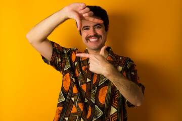 Positive young Caucasian man wearing printed shirt against yellow wall with cheerful expression, has good mood, gestures finger frame actively at camera.