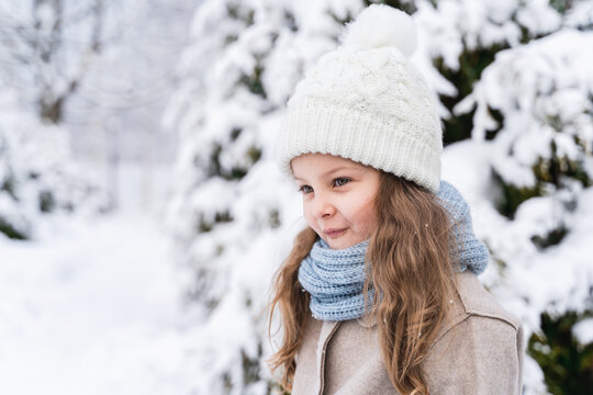 Little beautiful stylish girl in winter hat, woolen coat, blue scarf, snood with long hair. Kid walking, playing in forest, park among trees covered with snow. Country house yard. Fashionable image