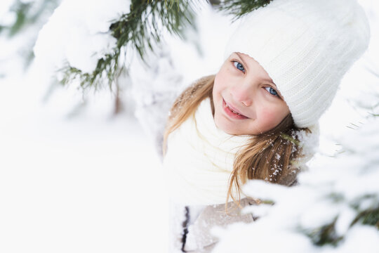Little beautiful stylish girl in winter hat, woolen coat, white scarf, snood with long hair. Kid walking, playing in forest, park among trees covered with snow. Country house yard. Fashionable image