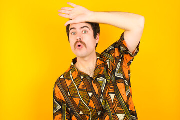 young Caucasian man wearing printed shirt against yellow wall wiping forehead with hand making phew gesture, expressing relief feels happy that he prevented huge disaster. It was close enough