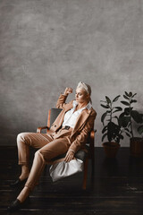 Young model woman in a trendy beige suit posing in a vintage armchair in minimalist interior