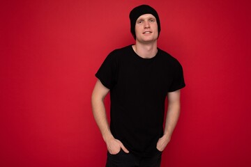 Shot of handsome happy smiling young man standing isolated over red background wall wearing black t-shirt for mockup and black hat and looking at camera