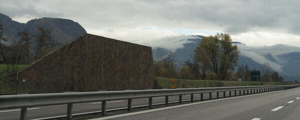 Double-tracked highway at the "Brenner" in Italy. Deciduous forest in a mountain landscape and a noise protection wall along the highway. Cloudy sky.