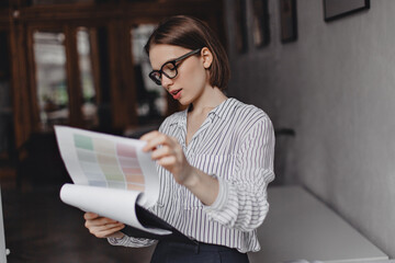 Focused business woman in white blouse is studying documents. Shot of girl with short hair wearing glasses and posing in office
