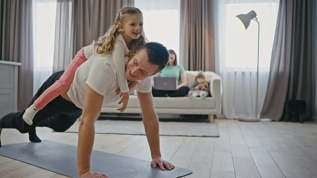Family fun. Strong dad practicing push ups with little daughter on his back, his wife and son on background