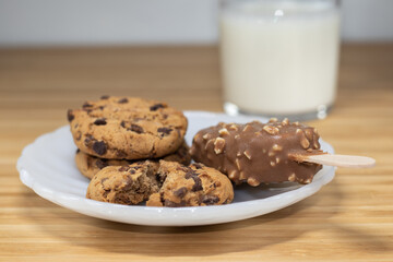 Chocolate chip cookies with a ice cream stick of a chocolate and almonds and a glass of milk in the brackground