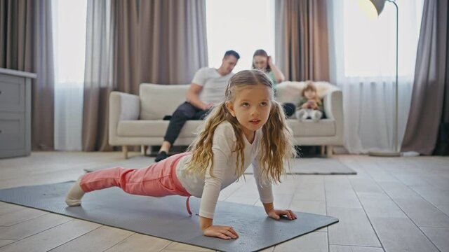 Gymnastics at home. Cute little girl stretching on fitness mat, her family watching her on background