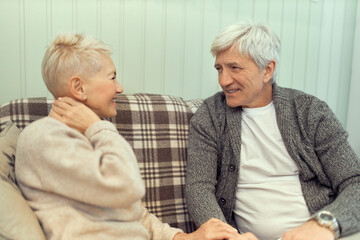 Adorable happy elderly couple relaxing indoors, sitting on couch, having fun. Stylish blonde female pensioner talking to her gray haired husband, sharing good positive news with him, laughing