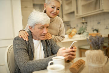 Fototapeta na wymiar Cozy image of senior man wearing cardigan sitting at dining table with cell phone in his hands, browsing internet together with his wife, watching pictures or making online order on web stores