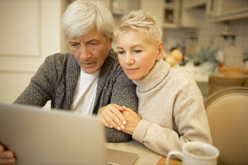 Serious elderly man and his blonde wife sitting in kitchen in front of open laptop, surfing internet together, signing up on website or social networks, looking at screen with concentrated expression